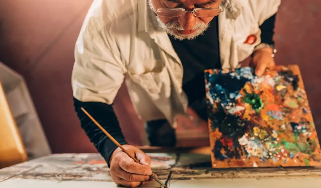 Older artist painting on a canvas in a dim room.