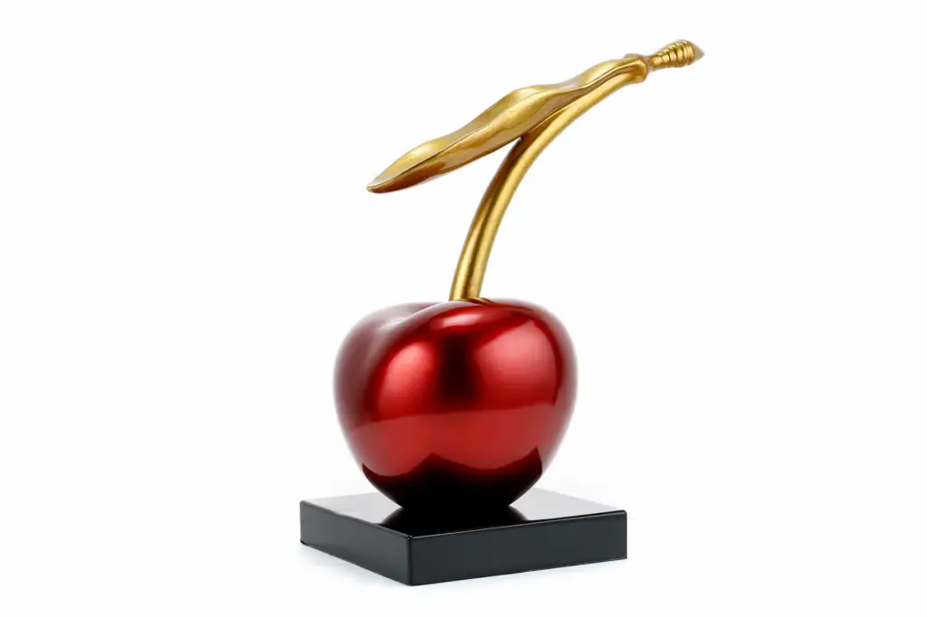 A contemporary sculpture of a giant, glossy red cherry with a long, elegant gold stem and leaf, sitting on a black square pedestal.